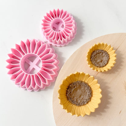 Two pink flower polymer clay cutters-shaped tools and two yellow flower-shaped molds on a white surface with a wooden board.