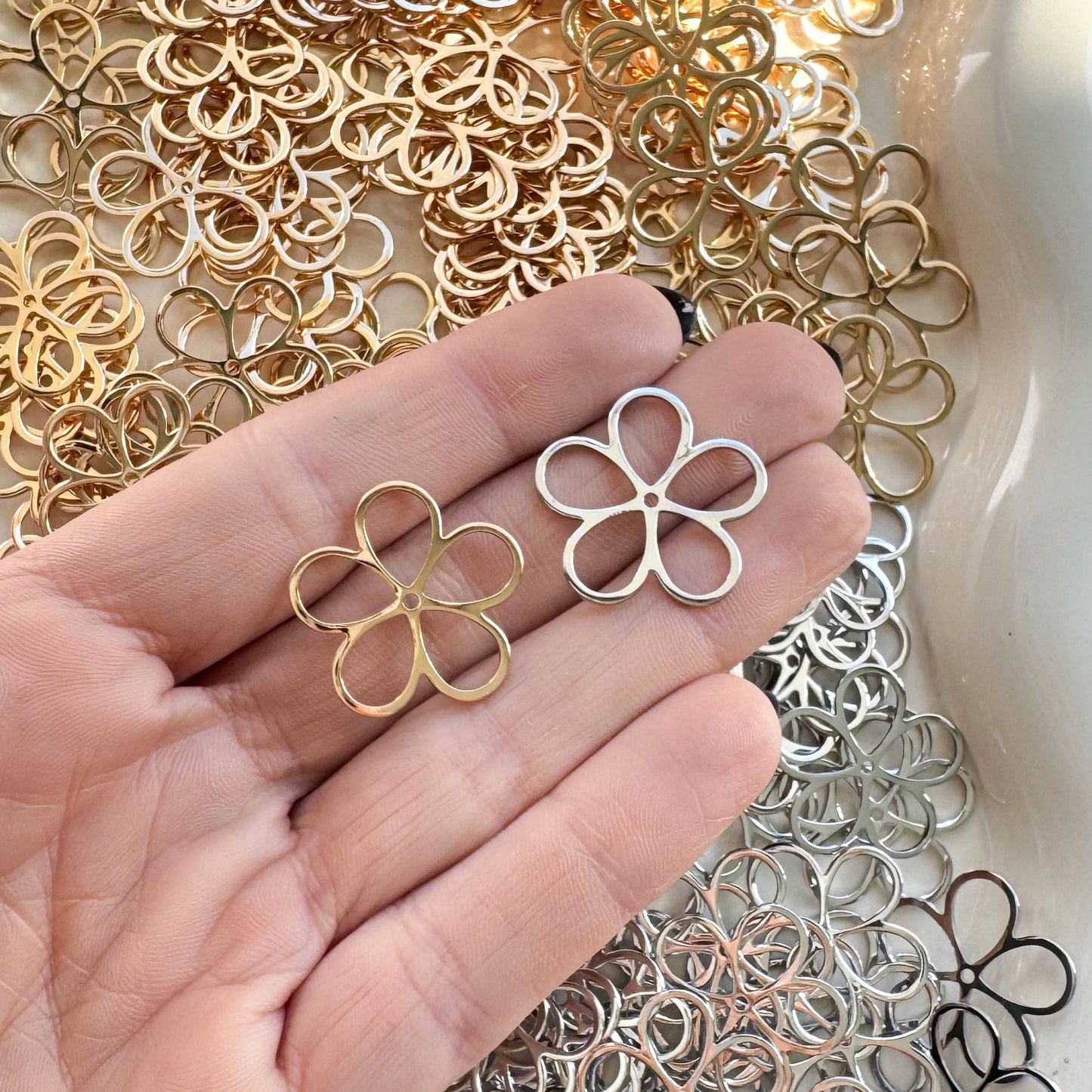 Hand holding a silver flower-shaped earring with a white dish filled with various metal flower-shaped jewelry pieces.