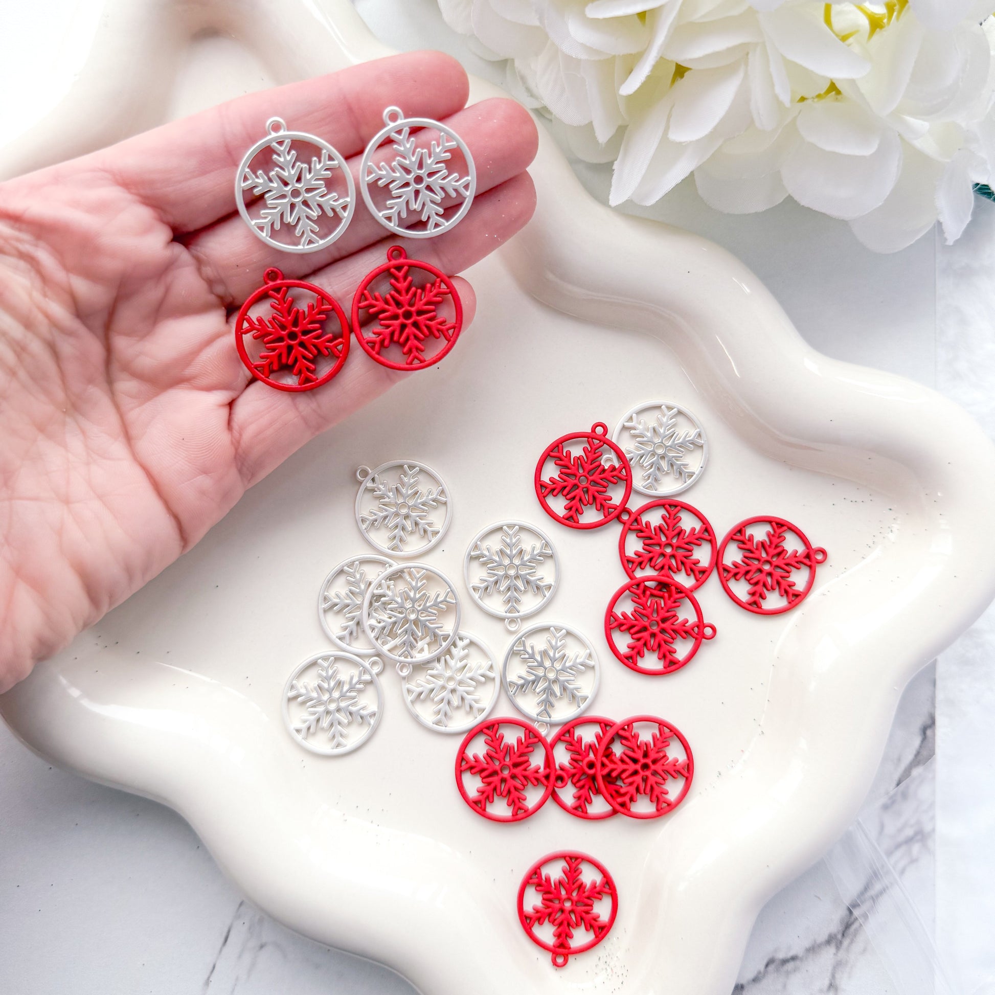 Red and silver earrings held by a hand with white flowers in the background